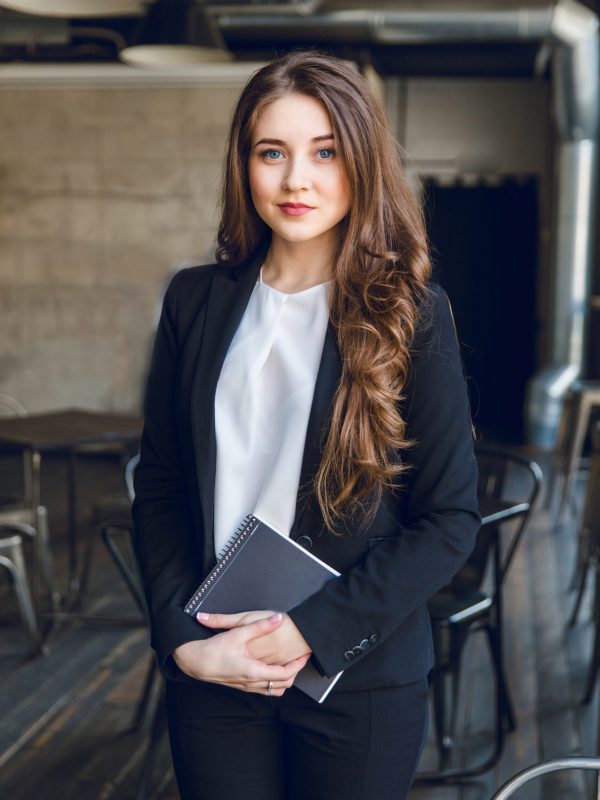 Brunette business woman with wavy long hair and blue eyes stands holding a notebook in hands. She wears a black suit and a white blouse. She is in a cafe with grey walls and dark furniture.
